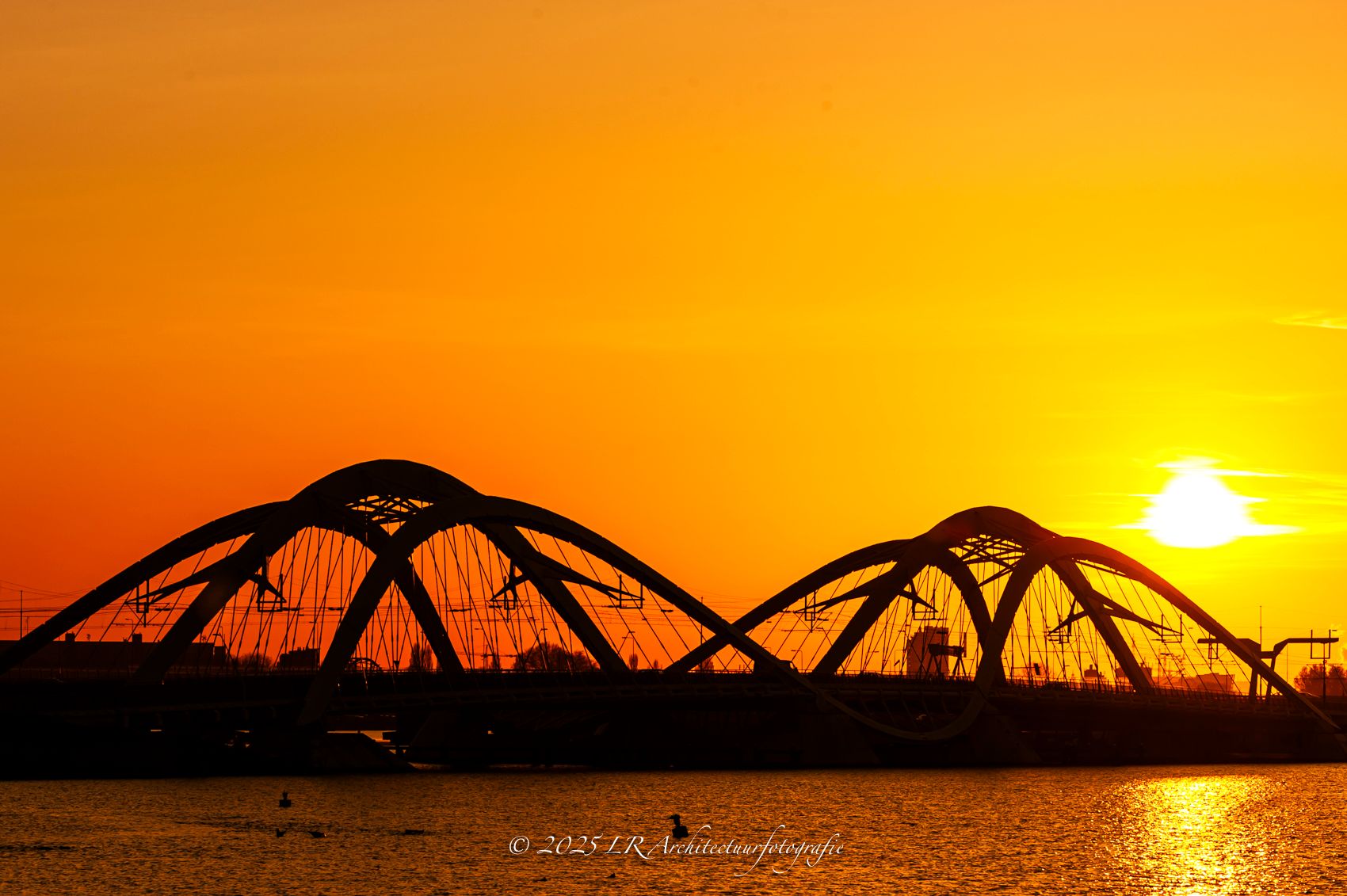 Het silhouet van een gebogen brugconstructie tegen een diep oranje avondlucht.