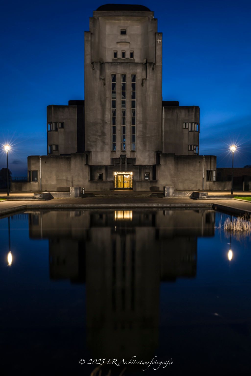 Radio Kootwijk in het blauwe uur, het monumentale betonnen zendgebouw, symmetrisch weerspiegeld in een stille vijver, met een warm verlichte entree.