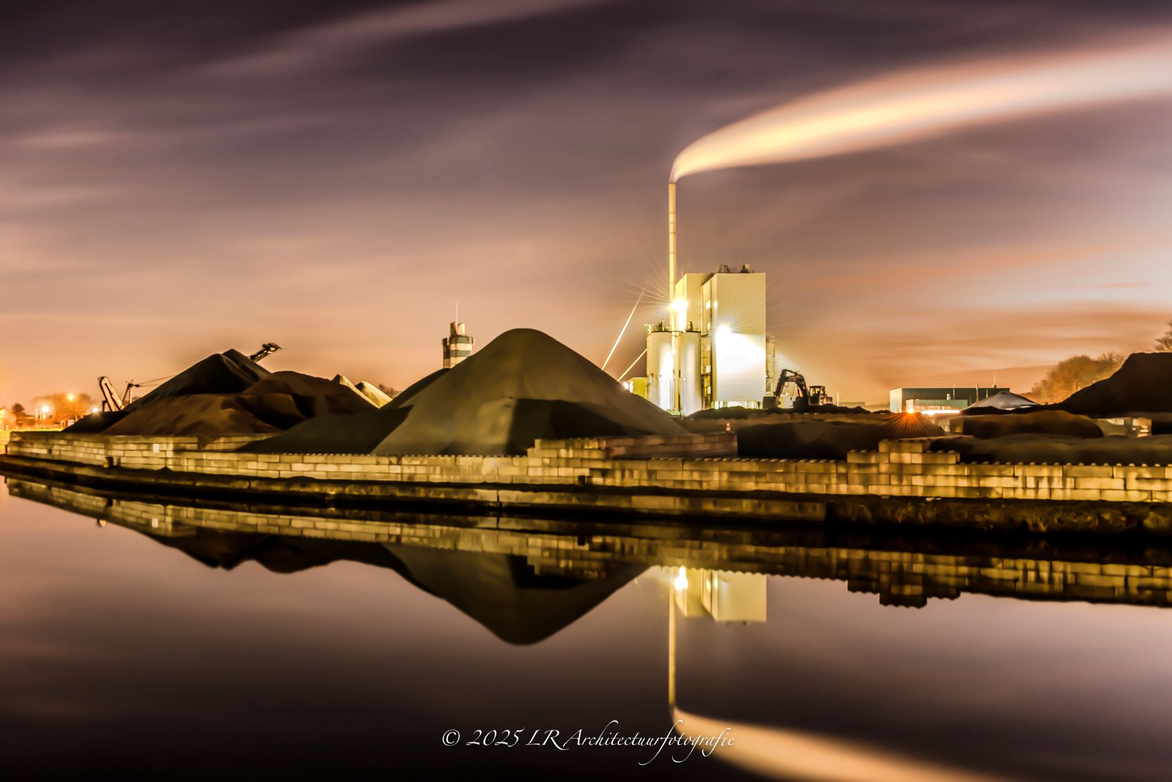 Een industrieel gebouw met warm verlichte ramen, gespiegeld in stil water in de avond.