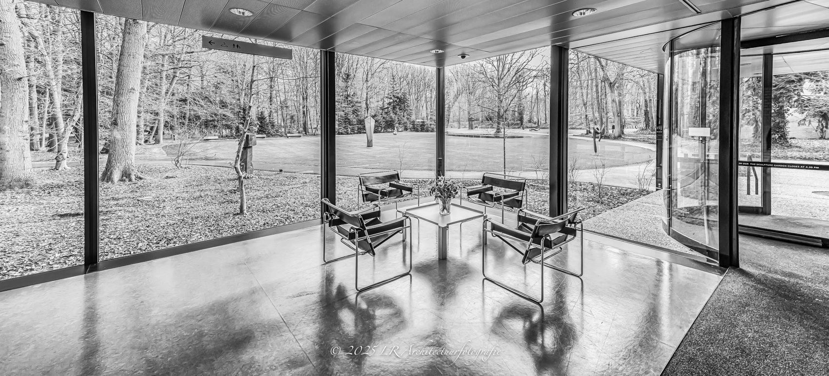 Interieur van een paviljoen met vier zwarte Wassily-fauteuils rond een lage tafel en glazen wanden die uitzicht bieden op de beeldentuin, in zwart-wit.
