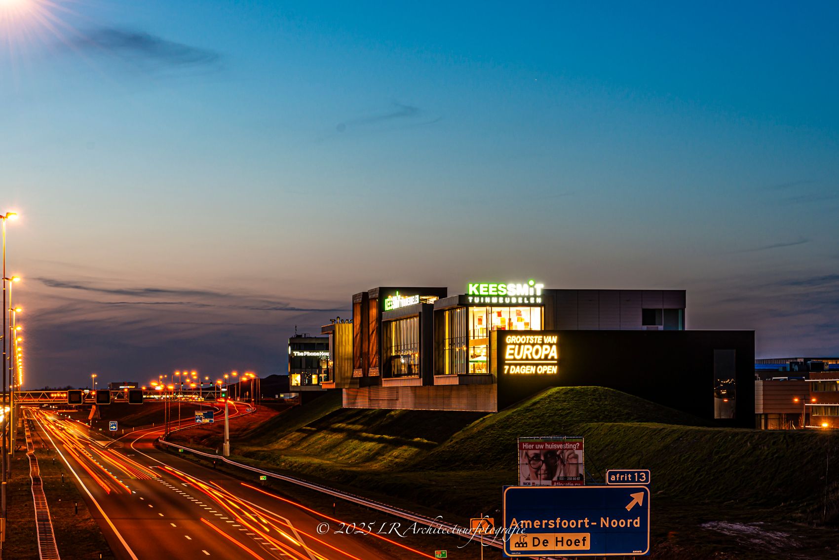 Een modern bedrijfsgebouw in de avond met een verlichte gevel, en lichtsporen van passerend verkeer op de weg ervoor.