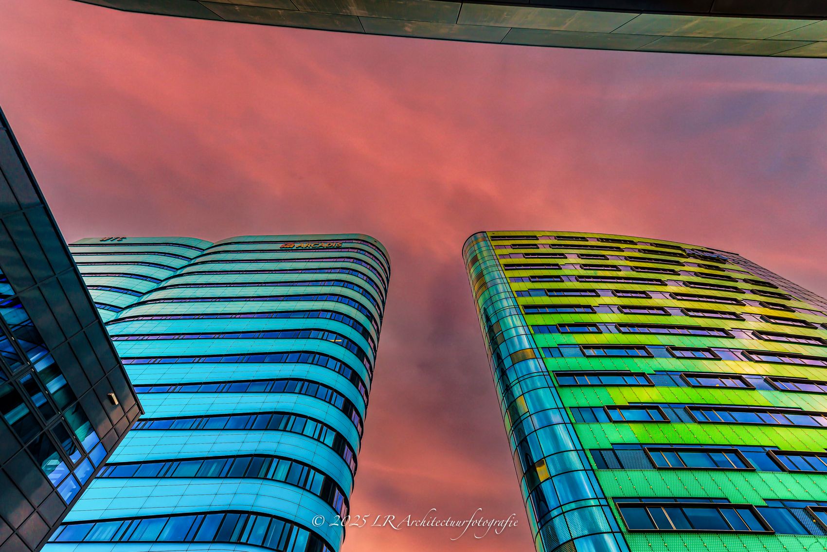 Twee kantoortorens met een golvende, kleurrijke gevel gefotografeerd van onderaf tegen een roze avondlucht in Arnhem.
