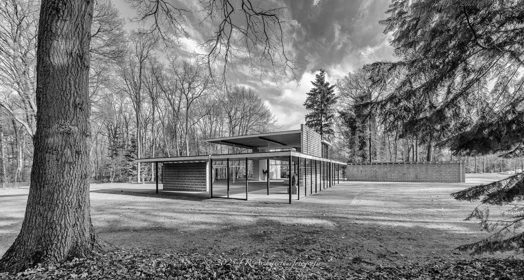 Buitenaanzicht van hetzelfde paviljoen tussen kale bomen, met een open overkapping en geperforeerde bakstenen wanden, in zwart-wit.