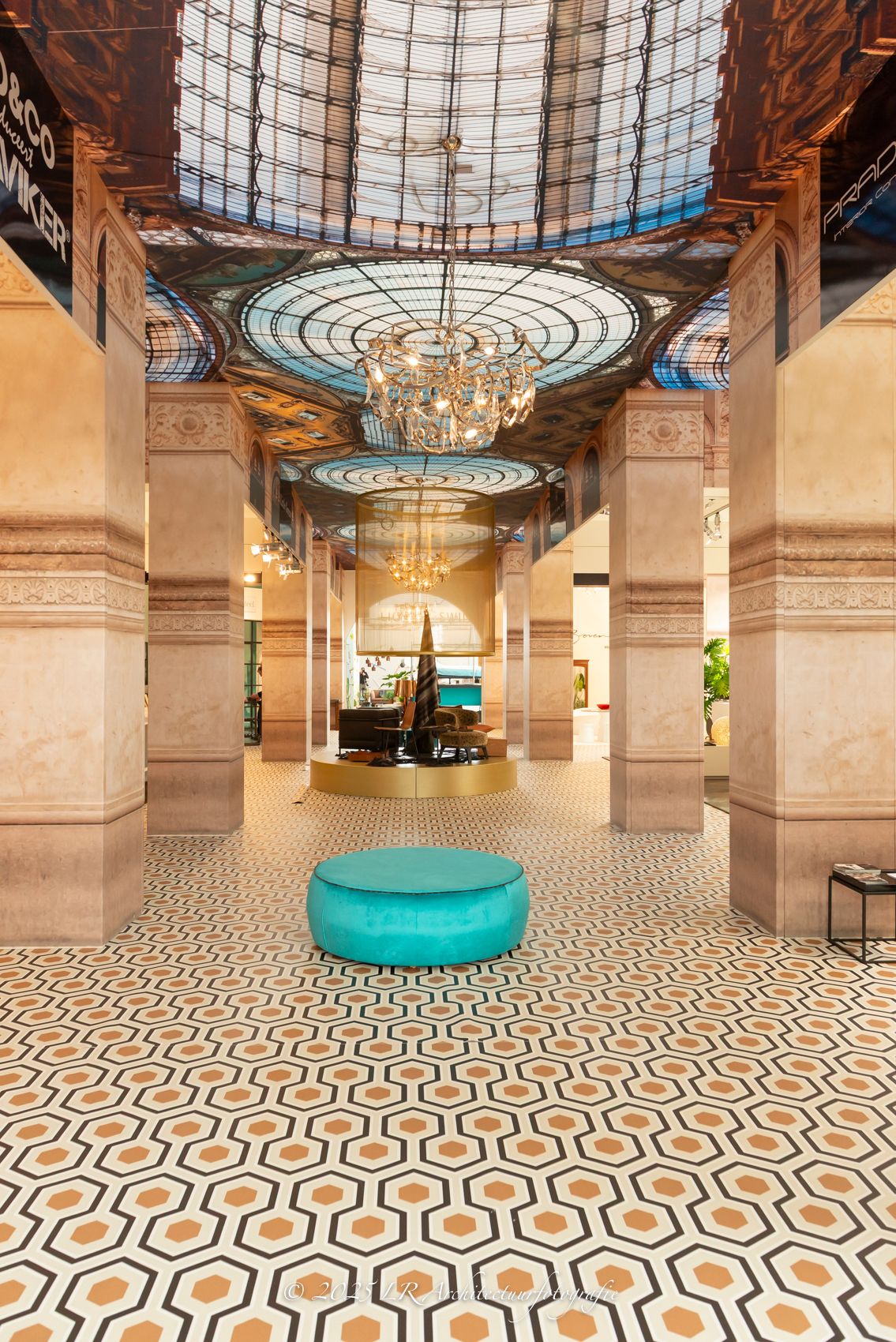 Een monumentale hal met marmeren zuilen, een glas-in-lood koepel en een kroonluchter, met een turquoise zitpoef op een geometrisch getegelde vloer.