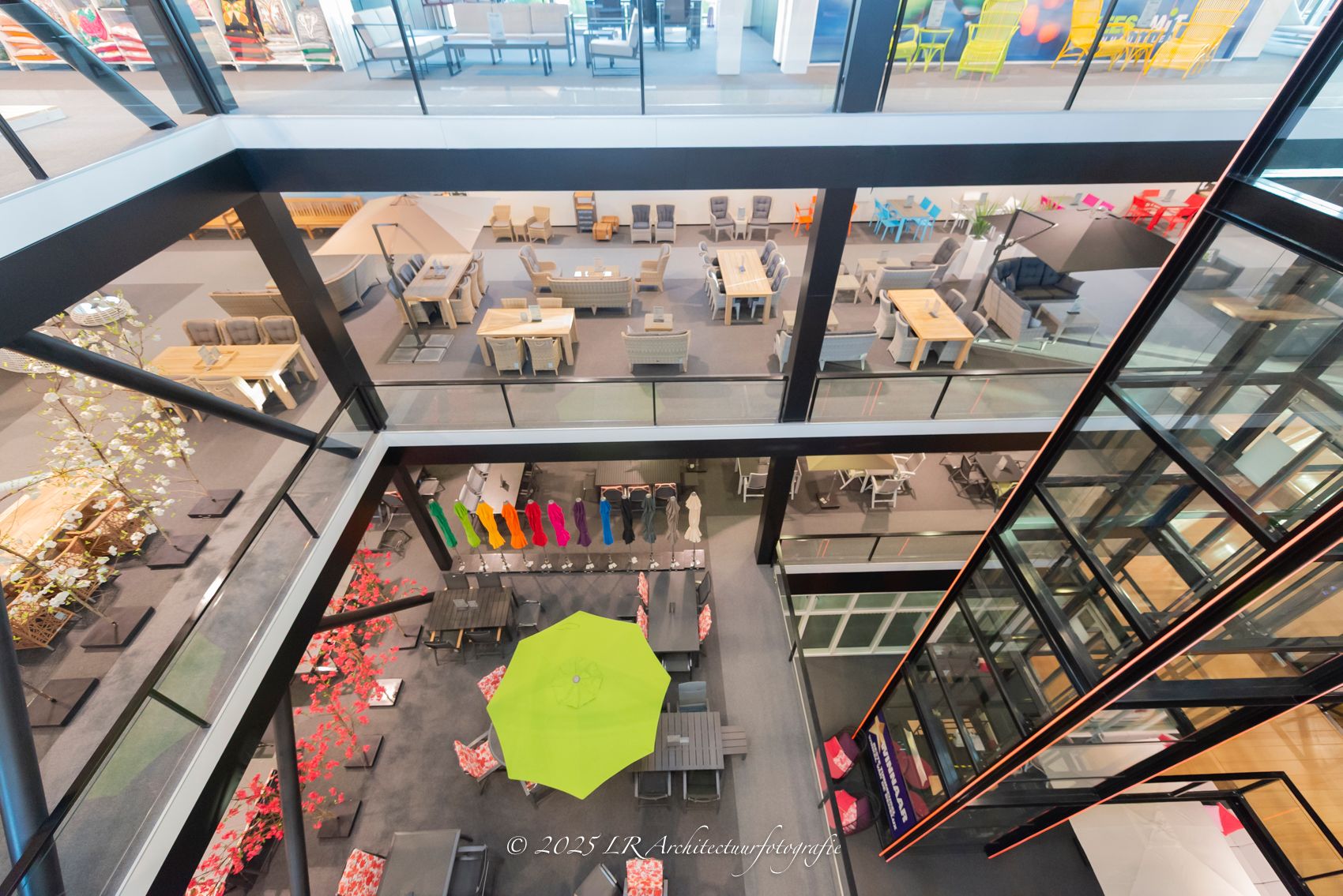 Een lichte showroom met een glazen vide, van bovenaf gefotografeerd, met tuinmeubelen en een opvallende gele parasol op de begane grond.
