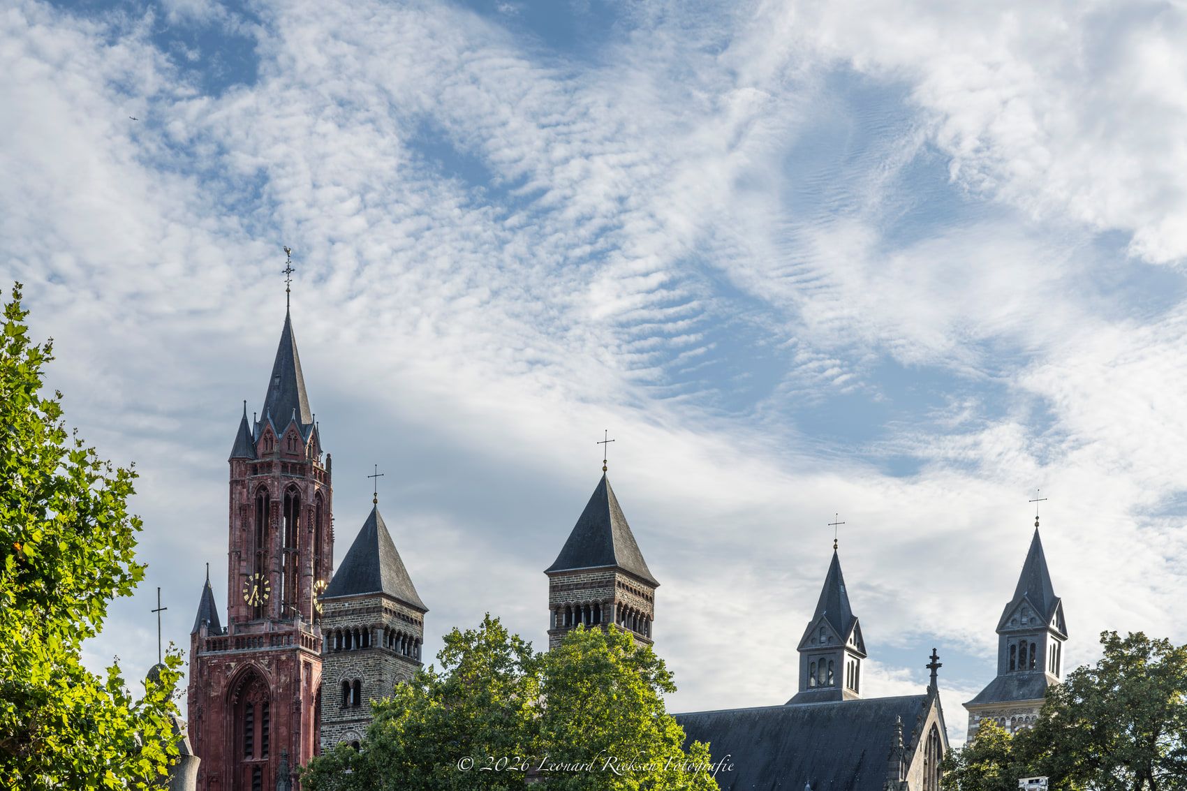 De rode toren van de Sint-Janskerk naast de grijze torens en het schip van de Sint-Servaasbasiliek, boven de boomkruinen aan het Vrijthof, onder een lucht met fijne schapenwolken.