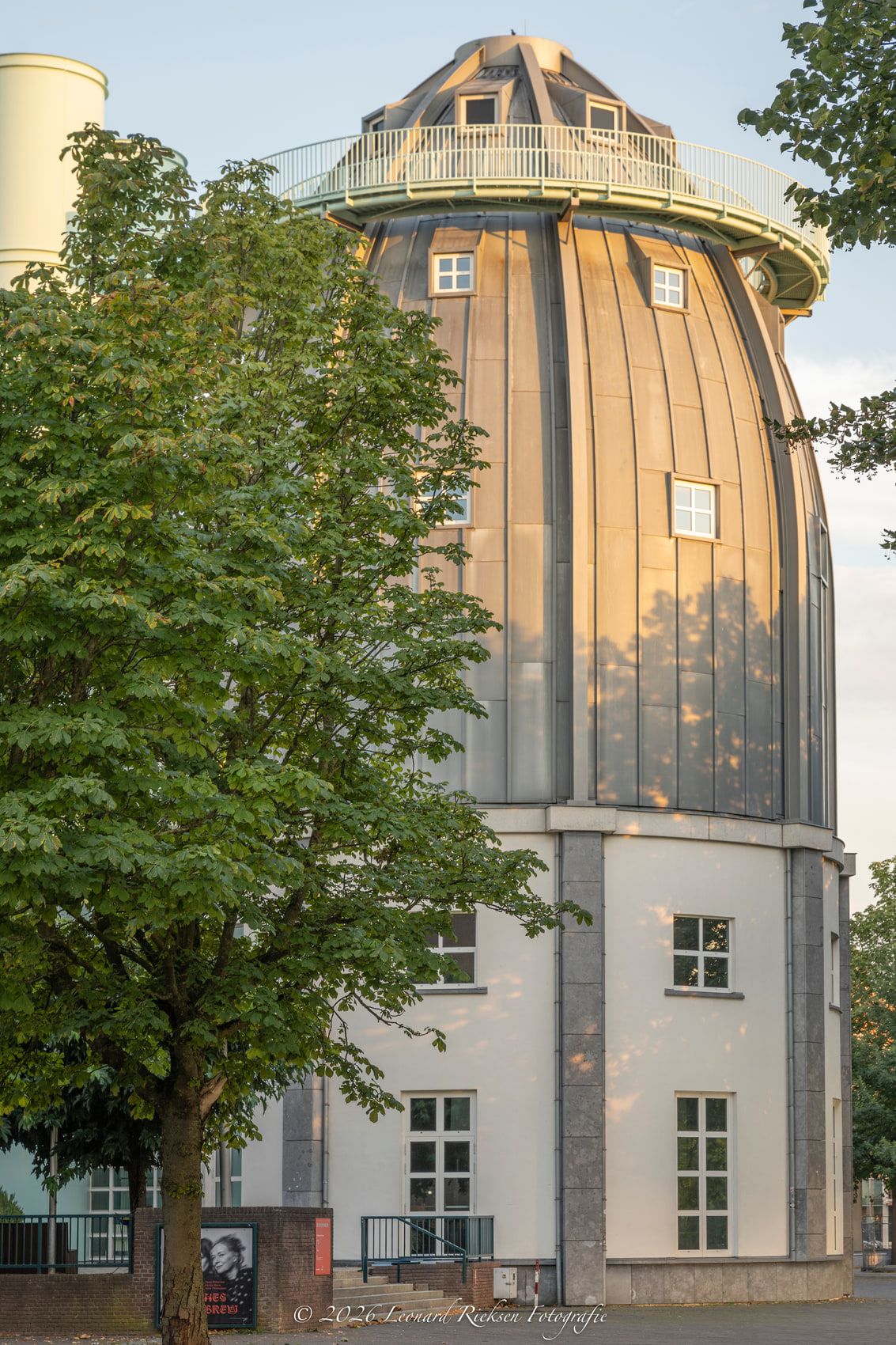 De met zink beklede koepeltoren van het Bonnefantenmuseum in laag avondlicht, half achter een kastanjeboom, met daaronder de lichte gevel en het balkon rond de toren.