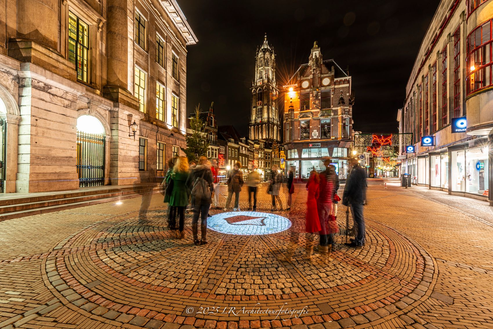 Een plein in de avond met wandelaars, waar verlichte historische gebouwen weerspiegelen in het natte plaveisel.