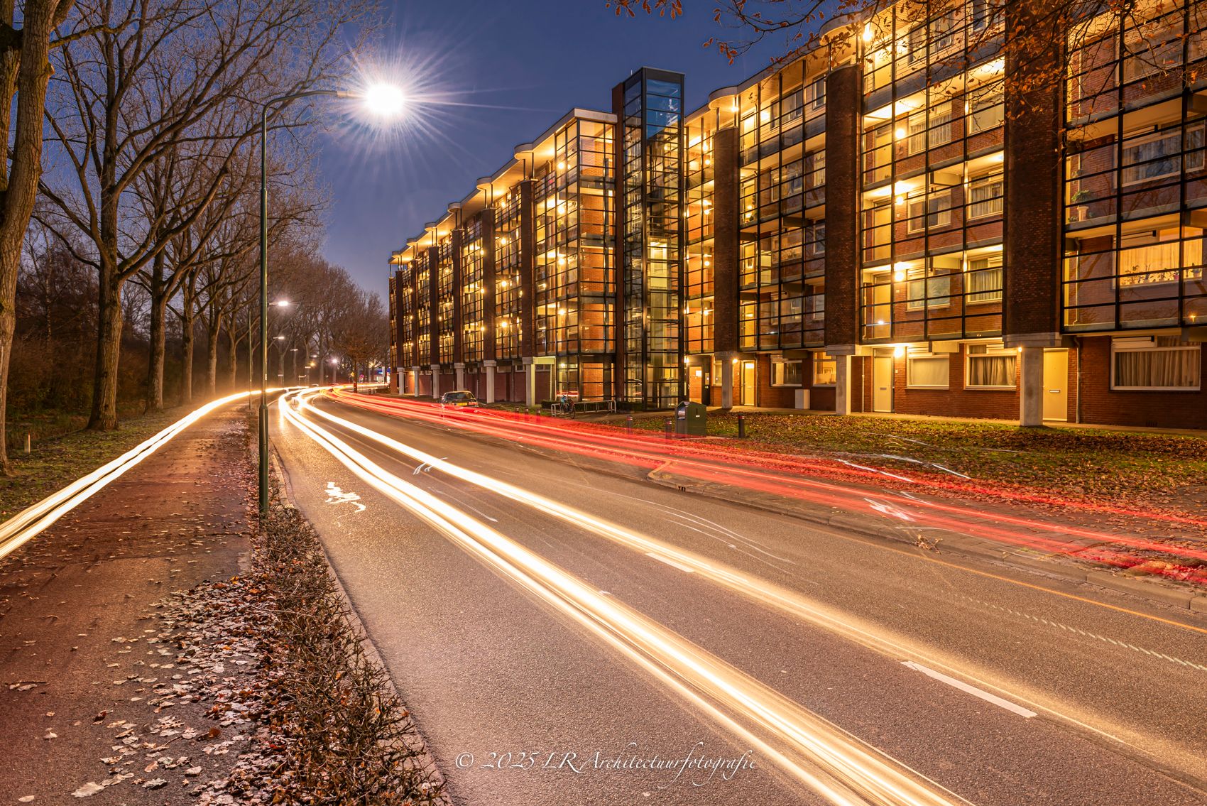 Lange sluitertijd langs een wooncomplex in de schemering: rode en witte lichtsporen van verkeer trekken door het beeld.