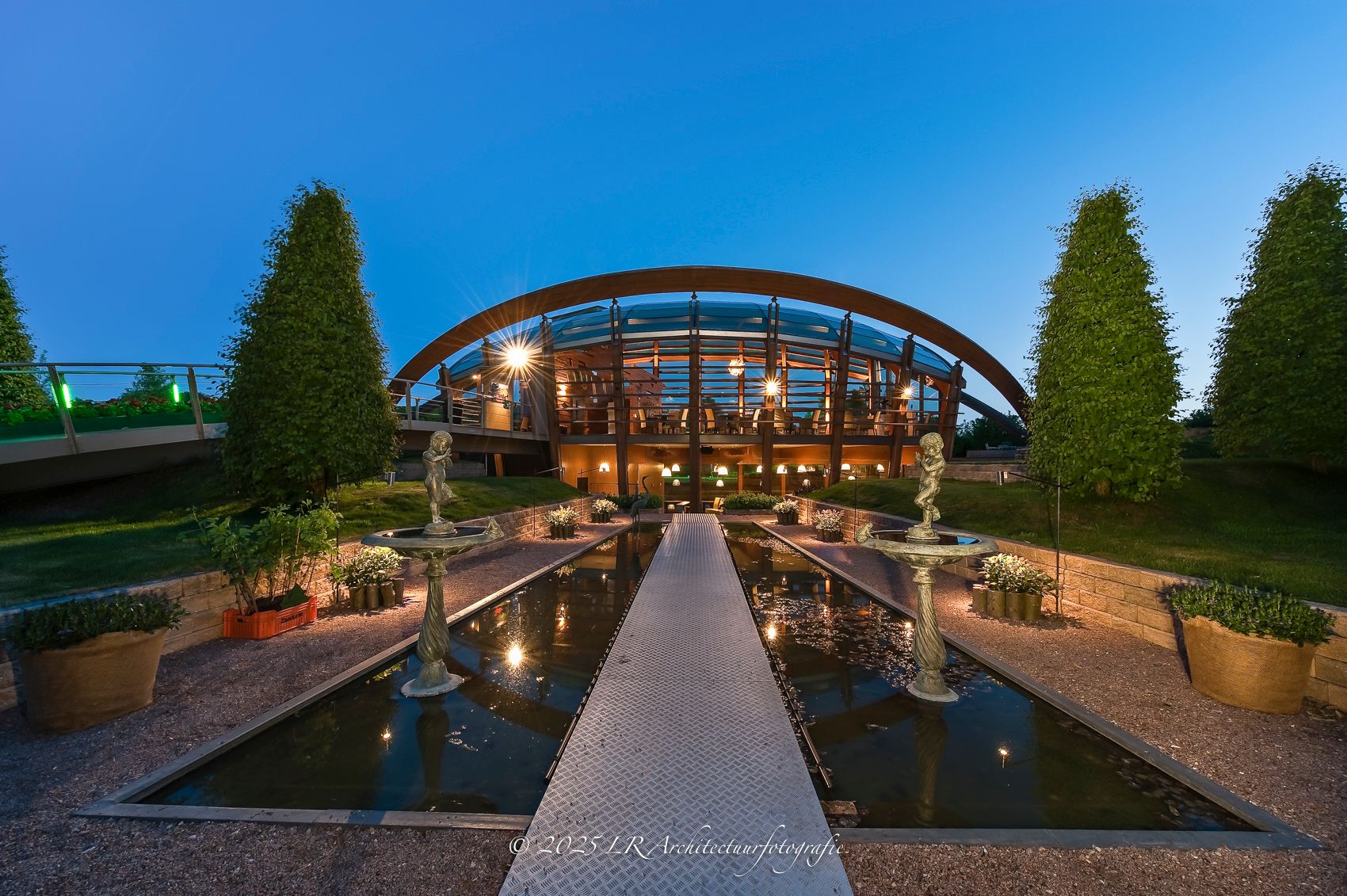 Een strak geometrisch glazen paviljoen boven stil water, met de constructie weerspiegeld in het wateroppervlak.